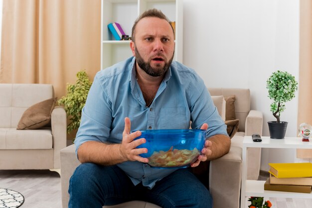 Шокированный взрослый славянский мужчина сидит на кресле с миской чипсов в гостиной