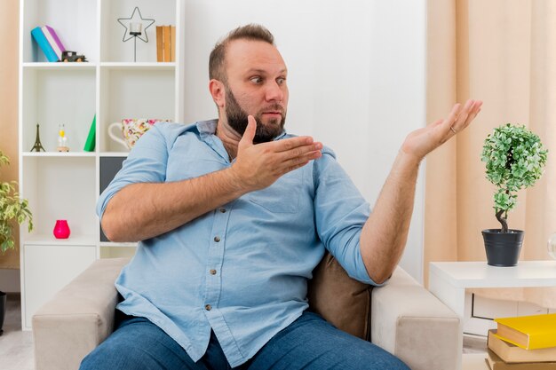 Shocked adult slavic man sits on armchair looking and pointing at side with two hands inside the living room