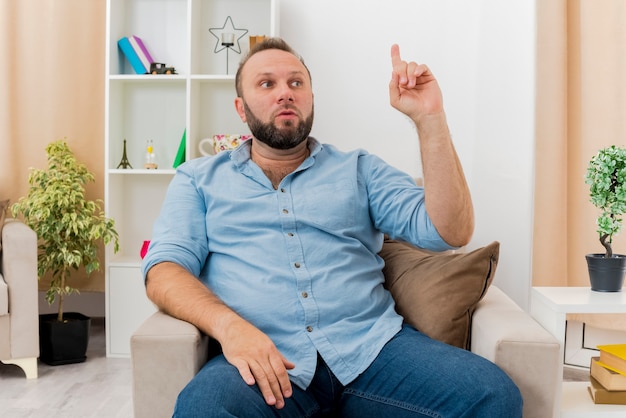 Shocked adult slavic man sits on armchair pointing up and looking at side inside the living room