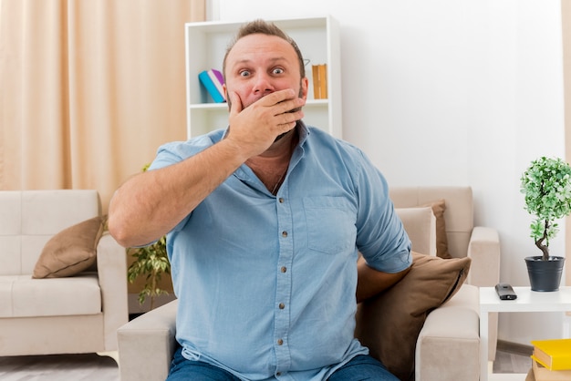 Shocked adult slavic man sits on armchair putting hand on mouth and holdign hand from behind inside the living room