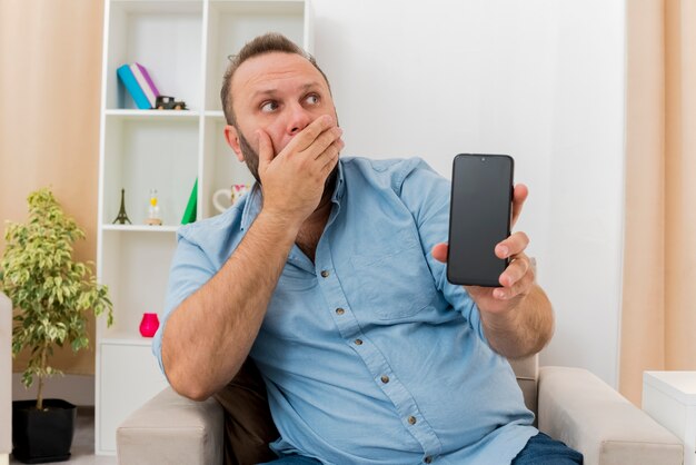 Shocked adult slavic man sits on armchair putting hand on mouth holding phone and looking at side inside the living room