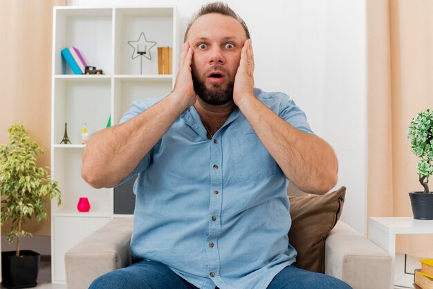 Shocked adult slavic man sits on armchair putting hands on face inside the living room