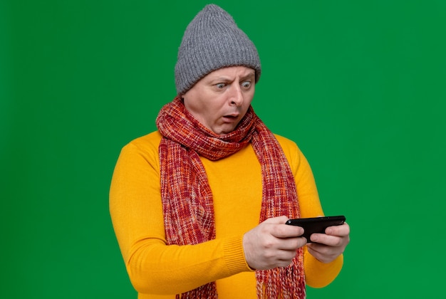 Шокированный взрослый славянский мужчина в зимней шапке и шарфе на шее держит и смотрит в телефон