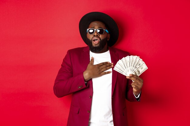 Shocked african american man holding hand on heart and gasping from excitement, showing huge amount of money, winning prize, standing over red background.