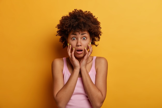 Free photo shocked african american woman gasps from wonder, touches face and stands worried, keeps jaw dropped and bugged eyes, cannot believe in bad news, poses over yellow wall. oh no, how terrible!