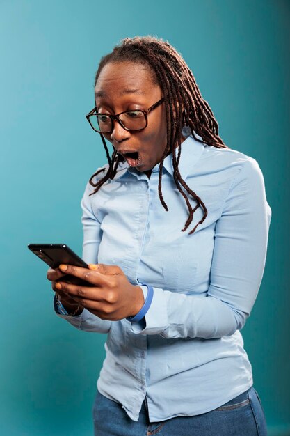 터치스크린 스마트폰 기기를 들고 충격을 받은 아프리카계 미국인 여성은 수신된 메시지에 놀랐습니다. 뉴스를 읽은 후 충격에 현대 전화와 함께 놀된 흥분된 젊은 성인 사람. 스튜디오 촬영