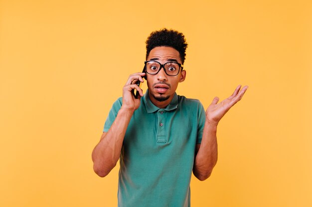電話で話している大きな眼鏡をかけたショックを受けたアフリカ人。スマートフォンでポーズをとる感情的な巻き毛の男の肖像画。
