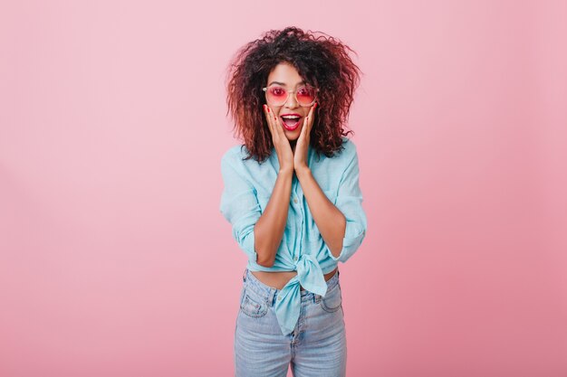 Shocked african lady with curly hairstyle expressing surprised emotions. Indoor portrait of amazing sporty black woman in denim pants touching her face.
