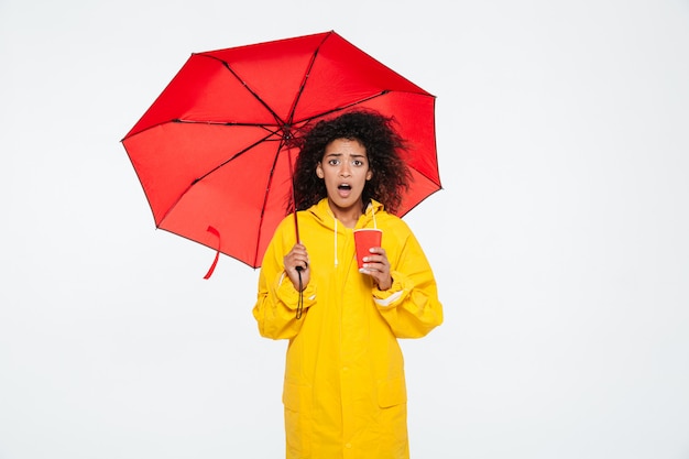 Shocked african woman in raincoat hiding under umbrella