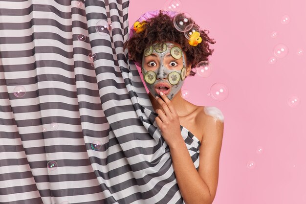 ショックを受けたアフリカ系アメリカ人の女性は、見つめられて目を大きく開いて、スキンケア用の美容マスクを適用し、ピンクの壁のシャボン玉の上にシャワーを浴びせます。