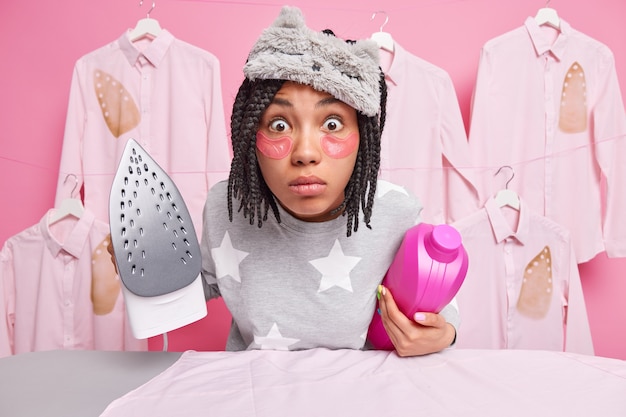 Shocked Afro American woman stares very surprised at camera busy ironing clothes after laundry holfd bottle of detergent and electric iron wears sleepmask soft pajama stays at home during quarantine