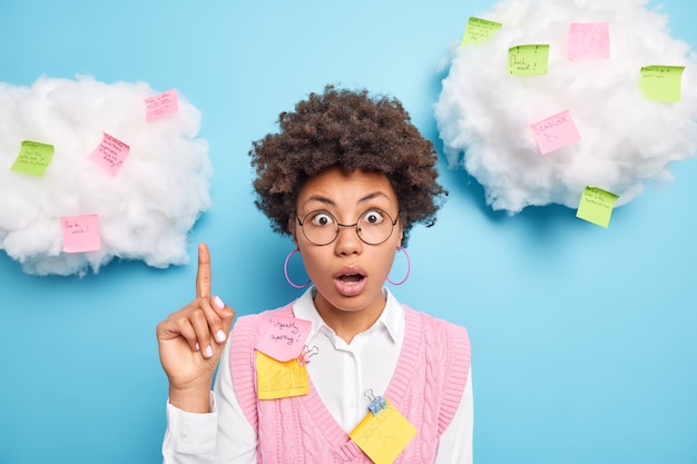 Shocked Afro American woman works in office works on marketing project points above with stunned expression on white clouds surrounded by colorful sticky notes