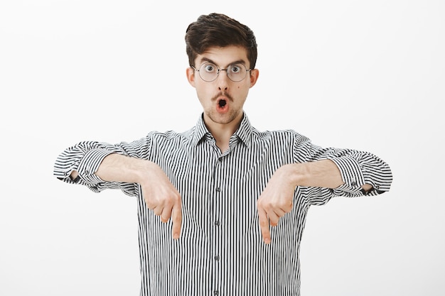 Free photo shocked amazed creative young guy with moustache in glasses, pointing down with index fingers, saying wow and dropping jaw, seeing something amazing and surprising downwards