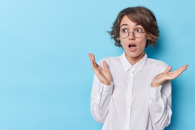 Shocked amazed woman with short hairstyle keeps mouth widely opened spreads palms reacts on unbelievable news wears round spectacles white shirt isolated on blue background copy space for your text