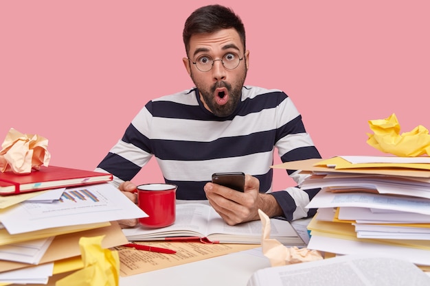 Free photo shocked amazed young caucasian man uses modern cell phone, drinks coffee, writes records in notepad, studies papers with graphics