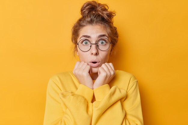 Shocked amazed young European woman gasps and keeps hands on cheeks stares surprised keeps mouth opened wears round spectacles for sight and casual jumper isolated over vivid yellow background