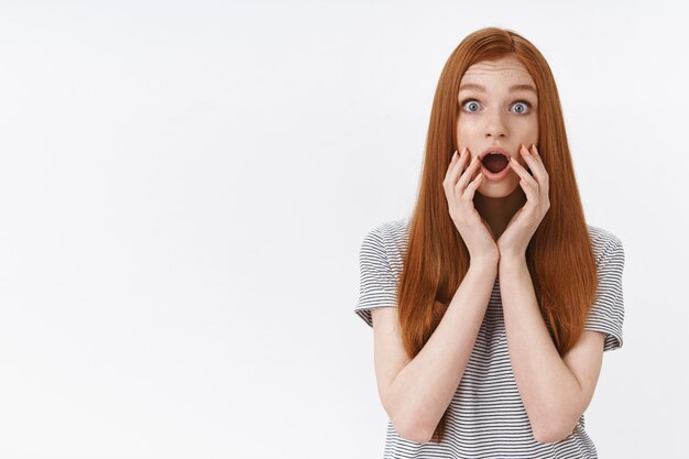 Shocked amused speechless ginger girl popping blue eyes drop jaw impressed touch face standing stunned surprised gossiping, reacting astonished hear disturbing news, white wall