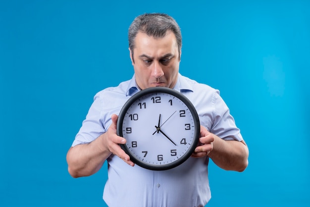 Shocked ans surprised middle age man in blue striped shirt holding wall clock with hands on a blue background