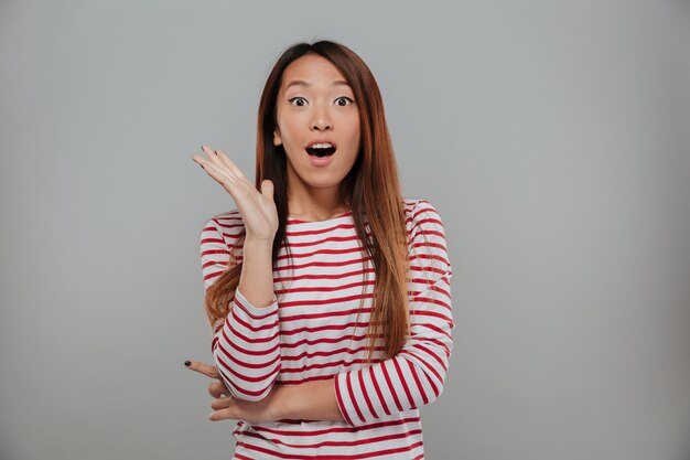 Shocked asian woman in sweater looking at the camera with open mouth over gray background