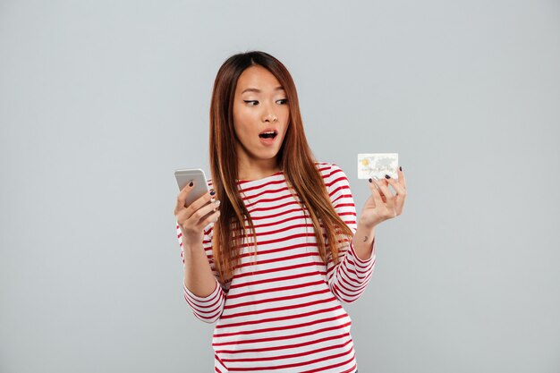 Shocked asian woman in sweater using smartphone and credit card over gray background