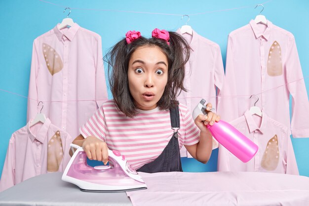 Shocked Asian woman with two pony tails holds detergent bottle busy ironing laundry at home