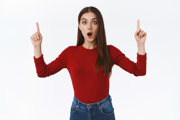 Shocked astonished brunette feminine girl in red sweater open mouth wondered and fascinated stare camera amazed look camera impressed as pointing up at stunning promo white background