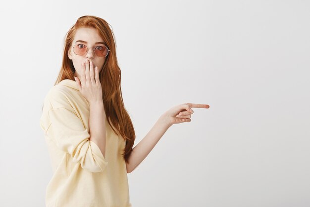Shocked and astounded redhead girl pointing finger right and gasping amazed