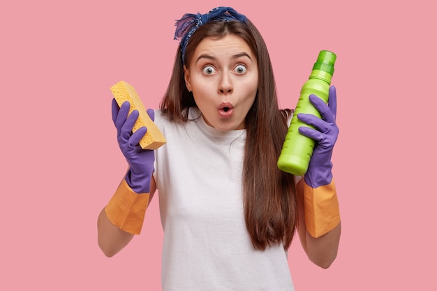 Shocked attractive young female from cleaning service, holds sponge and washing detergent, keeps jaw dropped