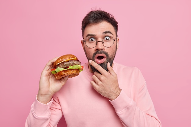 Shocked bearded adult man holds delicious hamburger eats fast food has unhealthy nutrition holds chin dressed in casual jumper 