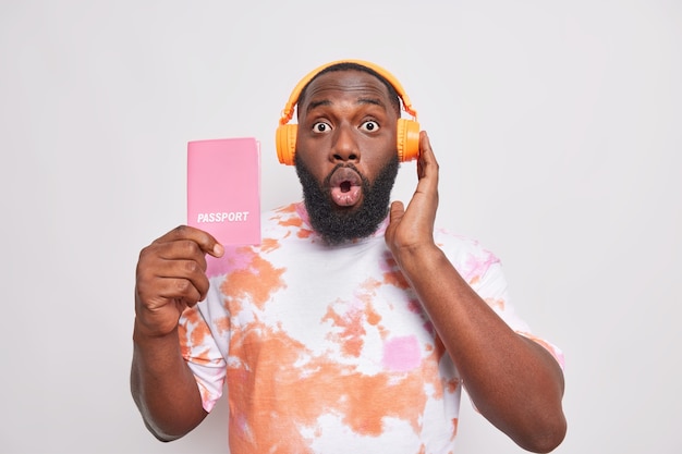 Shocked bearded adult man listens music before flight shows passport ready for trip finds out amazing news dressed in casual t shirt isolated over white wall got refusal of getting visa