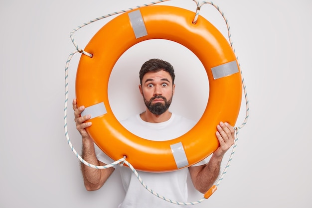 Потрясенный бородатый взрослый мужчина смотрит через надутый спасательный круг, смотрит в глаза, учится плавать или спасать людей, носит повседневную футболку, изолированную на белом фоне. Мужчина-спасатель позирует в помещении