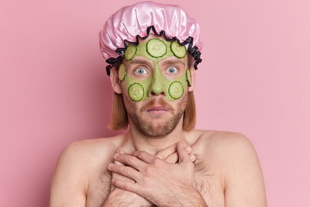 Shocked bearded European man stares bugged eyes keeps hands on chest applies green nourishing mask with cucumber slices wears bath hat.