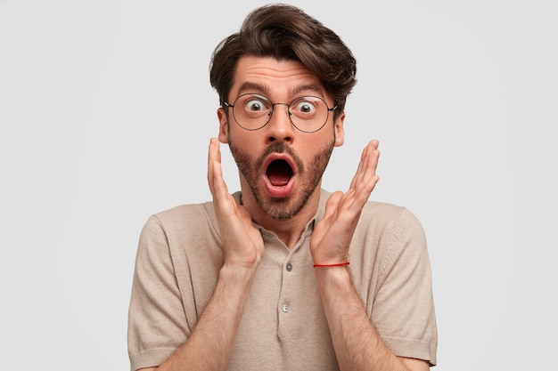 Shocked bearded man recieves unexpected news from friend, clasps hands near face, opens mouth widely, expresses surprisement, isolated on white wall