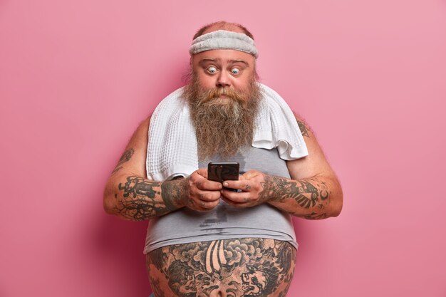 Shocked bearded man in sport outfit, takes break after physical exercises at home, holds smart phone, burns calories and fat, downloads application, poses against rosy wall. Thick fat guy with gadget