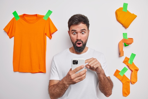 Shocked bearded man stares at smartphone screen surprised to read amazing news or someone tried to hack his phone dressed casually poses over white background with plastered orange items of clothing