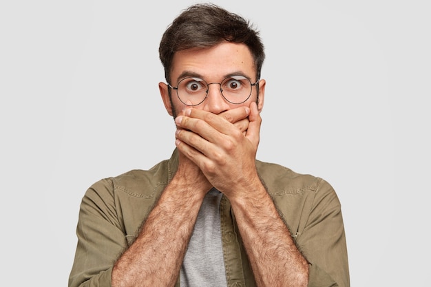 Shocked bearded young guy covers mouth with hands, has frightened expression, stares with eyes popped out, being speechless and mute, isolated over white wall. People, reaction concept