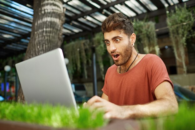 Потрясенный бородатый молодой человек с темными волосами и выпученными глазами, с открытым ртом, одетый в красную футболку, смотрит на экран своего ноутбука