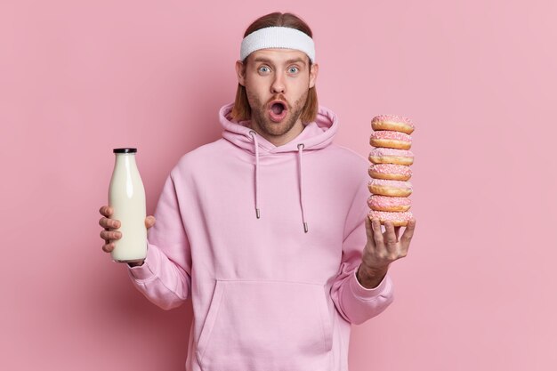 Shocked bearded young sportsman has temptation to eat junk food stares with widely opened mouth. Sporty guy wears hoodie and headband holds glass bottle of milk and many sweet donuts