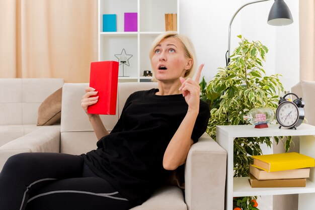 Shocked beautiful blonde russian woman lying on armchair holding book looking and pointing up inside the living room