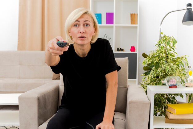 Shocked beautiful blonde russian woman sits on armchair holding tv remote looking forward inside the living room