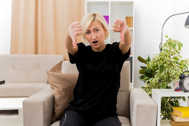 Shocked beautiful blonde russian woman sits on armchair thumbs down with two hands