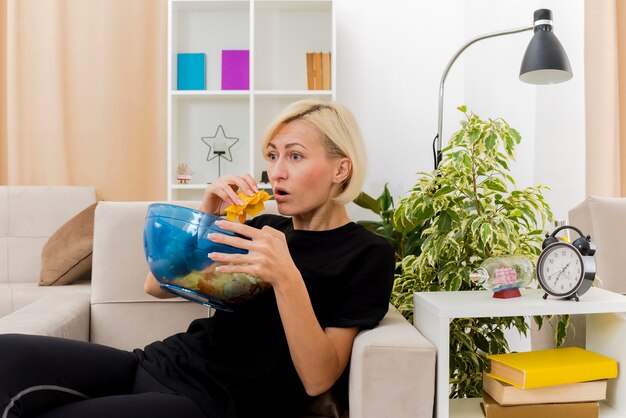 Shocked beautiful blonde woman lying on armchair holding and eating bowl of chips