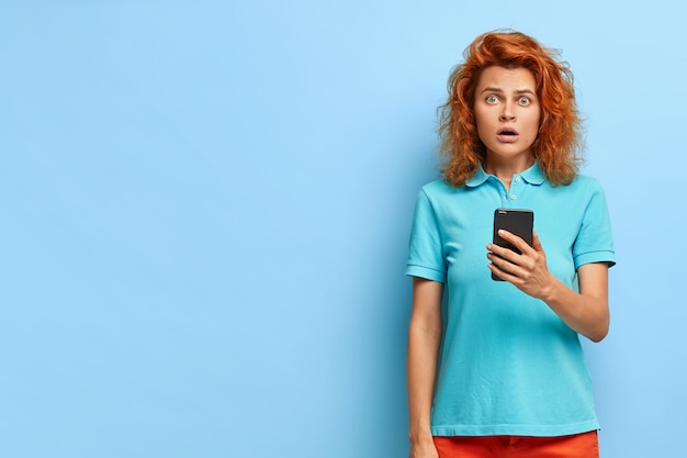 Шокированная красивая европейская женщина с рыжими волосами имеет впечатляющее выражение лица, держит современный мобильный телефон, получает уведомление, одета в повседневную одежду, модели у синей стены с местом для текста.