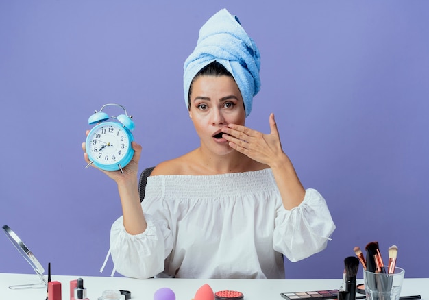 Shocked beautiful girl wrapped hair towel sits at table with makeup tools holding alarm clock and puts hand on mouth looking isolated on purple wall