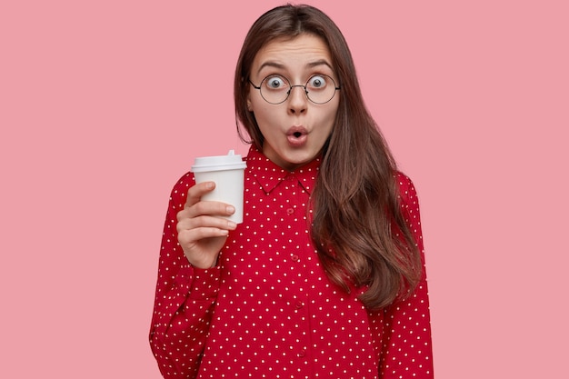 Shocked beautiful woman holds takeaway coffee, opens mouth, surprised to hear rumors