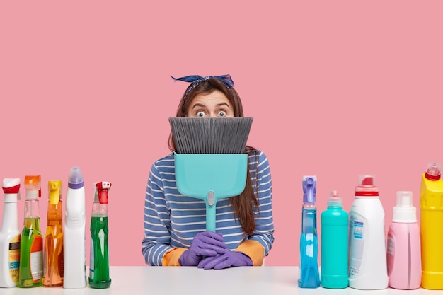 Shocked beautiful woman hostess hides behind broom, has bugged eyes, surprised with dirty untidy house