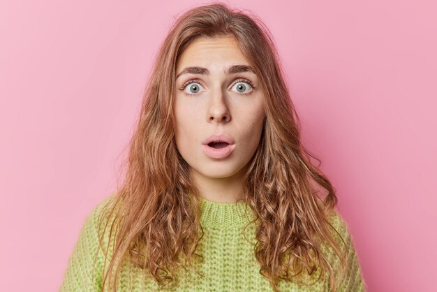 Shocked beautiful young long haired European woman stares bugged eyes keeps mouth opened reacts on amazing news wears green knitted sweater isolated over pink background. Human reactions concept