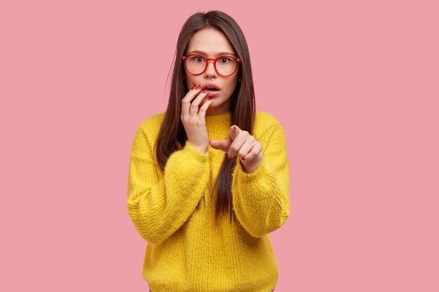 Shocked beautiful young woman with scared expression points at you, has worried frustrated look, dressed in yellow outfit