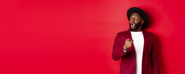 Shocked black man gasping amazed and pointing finger at camera recognize someone standing in red blazer and hat against studio background