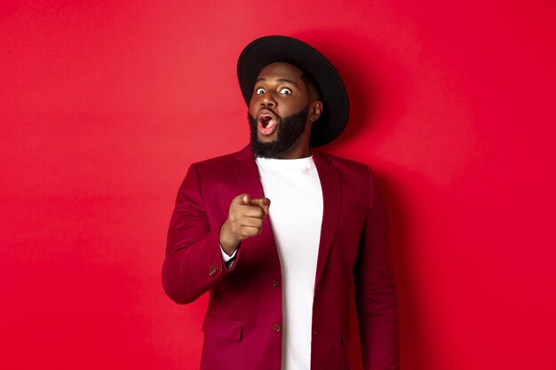 Shocked Black man gasping amazed and pointing finger at camera, recognize someone, standing in red blazer and hat against studio background.